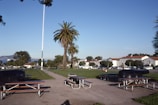 A park setting with picnic tables and benches on a paved area surrounded by green grass. Tall palm trees stand prominently against a clear blue sky. In the background, there are buildings with white walls and red roofs, and a parking lot with several vehicles. Distant hills and a glimpse of water can be seen in the far distance.