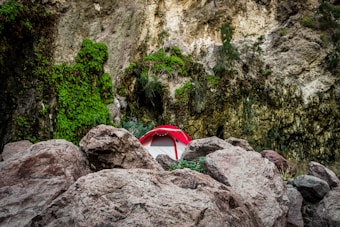 A vibrant red tent is nestled among large rugged boulders, surrounded by towering vertical rock faces covered in lush green vegetation. The scene conveys a sense of rugged outdoor adventure amid natural rock formations.