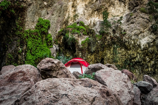 A vibrant red tent is nestled among large rugged boulders, surrounded by towering vertical rock faces covered in lush green vegetation. The scene conveys a sense of rugged outdoor adventure amid natural rock formations.