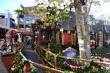 A festive street scene decorated for the holidays, featuring greenery, red bows, and ornaments adorning a walkway leading to a cozy-looking building. A sign for the Children's Hospital Los Angeles is prominently displayed. The area is surrounded by trees wrapped in lights, and there are people walking nearby. Storefronts and holiday figurines add to the vibrant seasonal atmosphere.
