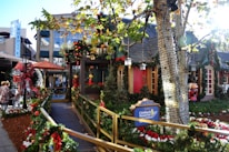 A festive street scene decorated for the holidays, featuring greenery, red bows, and ornaments adorning a walkway leading to a cozy-looking building. A sign for the Children's Hospital Los Angeles is prominently displayed. The area is surrounded by trees wrapped in lights, and there are people walking nearby. Storefronts and holiday figurines add to the vibrant seasonal atmosphere.