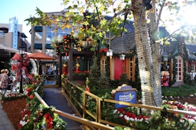 A festive street scene decorated for the holidays, featuring greenery, red bows, and ornaments adorning a walkway leading to a cozy-looking building. A sign for the Children's Hospital Los Angeles is prominently displayed. The area is surrounded by trees wrapped in lights, and there are people walking nearby. Storefronts and holiday figurines add to the vibrant seasonal atmosphere.