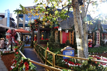 A festive street scene decorated for the holidays, featuring greenery, red bows, and ornaments adorning a walkway leading to a cozy-looking building. A sign for the Children's Hospital Los Angeles is prominently displayed. The area is surrounded by trees wrapped in lights, and there are people walking nearby. Storefronts and holiday figurines add to the vibrant seasonal atmosphere.