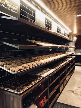 A bakery display with several shelves filled with a variety of donuts. The top shelf has donuts topped with nuts, the middle shelf contains assorted plain and sprinkled donuts, while the bottom shelf holds chocolate-glazed donuts. The background features dark tiled walls and a wooden ceiling, with a soft light illuminating the baked goods.