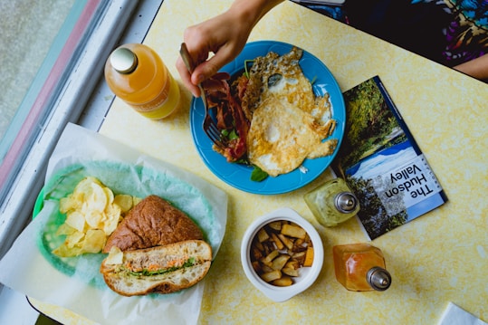 A table setting featuring a hand reaching for a blue plate containing crispy bacon and fried eggs. Adjacent to the plate are a bottle of juice, a glass salt shaker, and a pepper shaker. A paper-lined tray holds a sandwich and potato chips. A bowl with diced fried potatoes and a brochure titled 'The Hudson Valley' are also present.