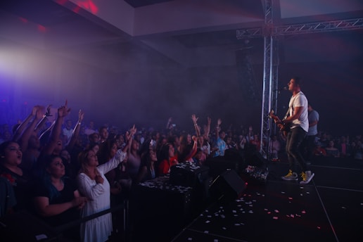 A lively concert scene with a band performing on stage. The lead singer and guitarist is enthusiastically engaging with the crowd, who are reaching out and cheering. The venue is filled with a smoke effect, enhancing the concert atmosphere, and bright stage lights create dynamic lighting.