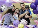 A family gathers closely, smiling and holding one another, surrounded by colorful balloons with paw print designs. The setting suggests a festive or celebratory occasion, possibly a birthday or special event.