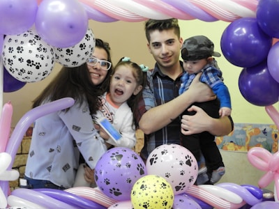 A family gathers closely, smiling and holding one another, surrounded by colorful balloons with paw print designs. The setting suggests a festive or celebratory occasion, possibly a birthday or special event.