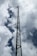A tall metal lattice tower stretches into the cloudy sky, equipped with several horizontal antennas. The structure is framed against a backdrop of white and gray clouds, with a glimpse of blue sky peeking through.