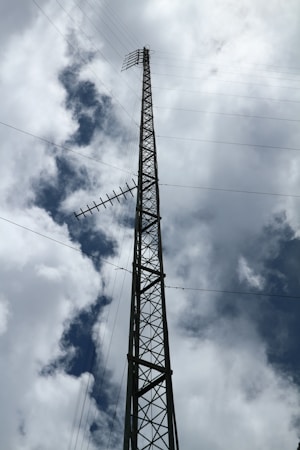 A tall metal lattice tower stretches into the cloudy sky, equipped with several horizontal antennas. The structure is framed against a backdrop of white and gray clouds, with a glimpse of blue sky peeking through.