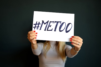 A person with long hair holds up a white sign featuring the handwritten hashtag '#MeToo' in bold black letters. The person wears a light long-sleeve top and has red-painted fingernails, standing against a plain dark background.
