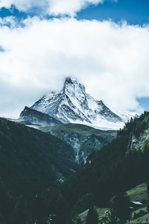 A majestic snow-covered mountain peak rises sharply against a backdrop of fluffy white clouds and a blue sky. The foreground features lush green forests and valleys, creating a striking contrast with the stark whiteness of the mountain.