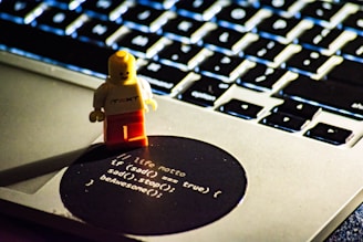 A small toy figure stands on the edge of a laptop keyboard, next to a sticker with a motivational programming quote. The background shows a close-up of the laptop keys, softly lit and partially out of focus.