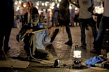 A bearded man, wearing a traditional hat and blue shirt, is sitting cross-legged on a paved area, playing a string instrument. The scene is dimly lit, with a lantern providing light in the foreground. There are a few objects on the ground around him, including a teapot and cups on a tray. In the background, a crowd of people can be seen, blurred and illuminated by strings of lights, suggesting a lively night market setting.