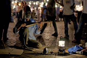 A bearded man, wearing a traditional hat and blue shirt, is sitting cross-legged on a paved area, playing a string instrument. The scene is dimly lit, with a lantern providing light in the foreground. There are a few objects on the ground around him, including a teapot and cups on a tray. In the background, a crowd of people can be seen, blurred and illuminated by strings of lights, suggesting a lively night market setting.