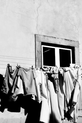 Clothes hang on a line strung across the exterior of a building, positioned below a single window. The wall appears to be weathered, and the garments, captured in monochrome, exhibit a range of textures and creases as they are draped over the line.