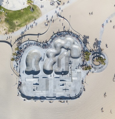 An aerial view captures a large concrete skatepark adjacent to a sandy beach. The park features smooth, curving bowls and ramps surrounded by numerous people casting long shadows. The layout includes paths and greenery nearby, along with palm trees and a promenade.