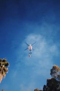 A helicopter is flying in the sky with a clear view from below. The helicopter is attached to a bucket that is often used for aerial firefighting. The background shows a blue sky with some scattered clouds. Tall trees with green foliage frame the image edges, giving a sense of height and open space.
