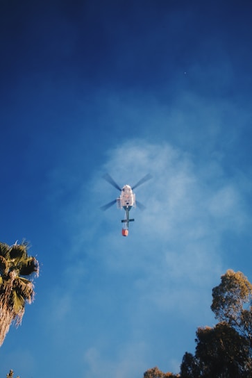 A helicopter is flying in the sky with a clear view from below. The helicopter is attached to a bucket that is often used for aerial firefighting. The background shows a blue sky with some scattered clouds. Tall trees with green foliage frame the image edges, giving a sense of height and open space.