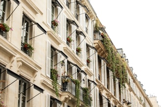 A row of elegant, classic European apartment buildings with beige stone facades, adorned with black wrought iron balconies. The balconies are decorated with abundant green plants and colorful flowers cascading down. White shutters frame the windows, providing a harmonious look.