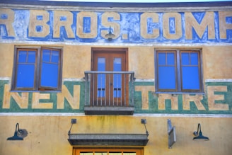 A vintage building facade featuring a blue and yellow sign with large, weathered letters. The upper section has two symmetrical windows with blue-tinted glass that reflect the sky, and a central wooden door with a small balcony. Below, there are industrial-style lamps on either side.