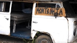A weathered, white van with a slightly rusty exterior has its side door open, revealing a makeshift sleeping area. Inside, there is a plaid mattress resting on a wooden frame. A cardboard sign attached to the vehicle reads 'Montañita.'