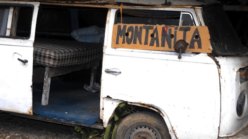 A weathered, white van with a slightly rusty exterior has its side door open, revealing a makeshift sleeping area. Inside, there is a plaid mattress resting on a wooden frame. A cardboard sign attached to the vehicle reads 'Montañita.'