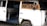 A weathered, white van with a slightly rusty exterior has its side door open, revealing a makeshift sleeping area. Inside, there is a plaid mattress resting on a wooden frame. A cardboard sign attached to the vehicle reads 'Montañita.'