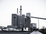 A large industrial plant with several cylindrical structures and tall smokestacks emitting steam or smoke. The building is surrounded by parked vehicles, including trucks, and is set against a clear sky. The scene feels somewhat desolate, with a concrete path leading to the complex.