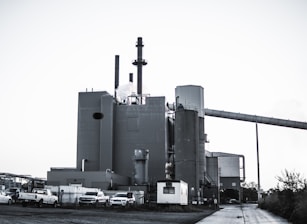 A large industrial plant with several cylindrical structures and tall smokestacks emitting steam or smoke. The building is surrounded by parked vehicles, including trucks, and is set against a clear sky. The scene feels somewhat desolate, with a concrete path leading to the complex.
