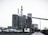 A large industrial plant with several cylindrical structures and tall smokestacks emitting steam or smoke. The building is surrounded by parked vehicles, including trucks, and is set against a clear sky. The scene feels somewhat desolate, with a concrete path leading to the complex.