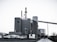 A large industrial plant with several cylindrical structures and tall smokestacks emitting steam or smoke. The building is surrounded by parked vehicles, including trucks, and is set against a clear sky. The scene feels somewhat desolate, with a concrete path leading to the complex.
