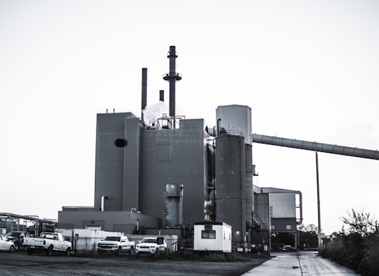 A large industrial plant with several cylindrical structures and tall smokestacks emitting steam or smoke. The building is surrounded by parked vehicles, including trucks, and is set against a clear sky. The scene feels somewhat desolate, with a concrete path leading to the complex.