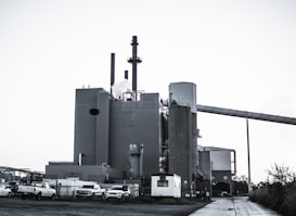 A large industrial plant with several cylindrical structures and tall smokestacks emitting steam or smoke. The building is surrounded by parked vehicles, including trucks, and is set against a clear sky. The scene feels somewhat desolate, with a concrete path leading to the complex.