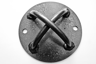 A close-up view of a round metal plate with two crossed metal rods attached to it. The plate has a textured surface and two circular holes on opposite sides. The grayscale image highlights the metallic texture and shadows.