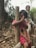 A young child in a pink dress is leaning against a wooden post, with hands clasped together near their face. In the background, another child is sitting on a log, surrounded by greenery and fallen leaves.