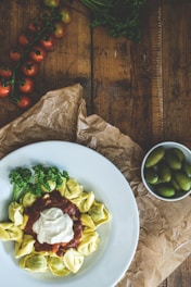 A wooden table displays a plate of tortellini served with a rich tomato sauce and a dollop of cream. Fresh parsley garnishes the pasta dish, adding a pop of green. In the background, a vine of ripe cherry tomatoes and a small white bowl filled with green olives are present. The rustic setting is complemented by crumpled brown paper.