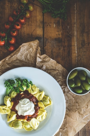 A wooden table displays a plate of tortellini served with a rich tomato sauce and a dollop of cream. Fresh parsley garnishes the pasta dish, adding a pop of green. In the background, a vine of ripe cherry tomatoes and a small white bowl filled with green olives are present. The rustic setting is complemented by crumpled brown paper.