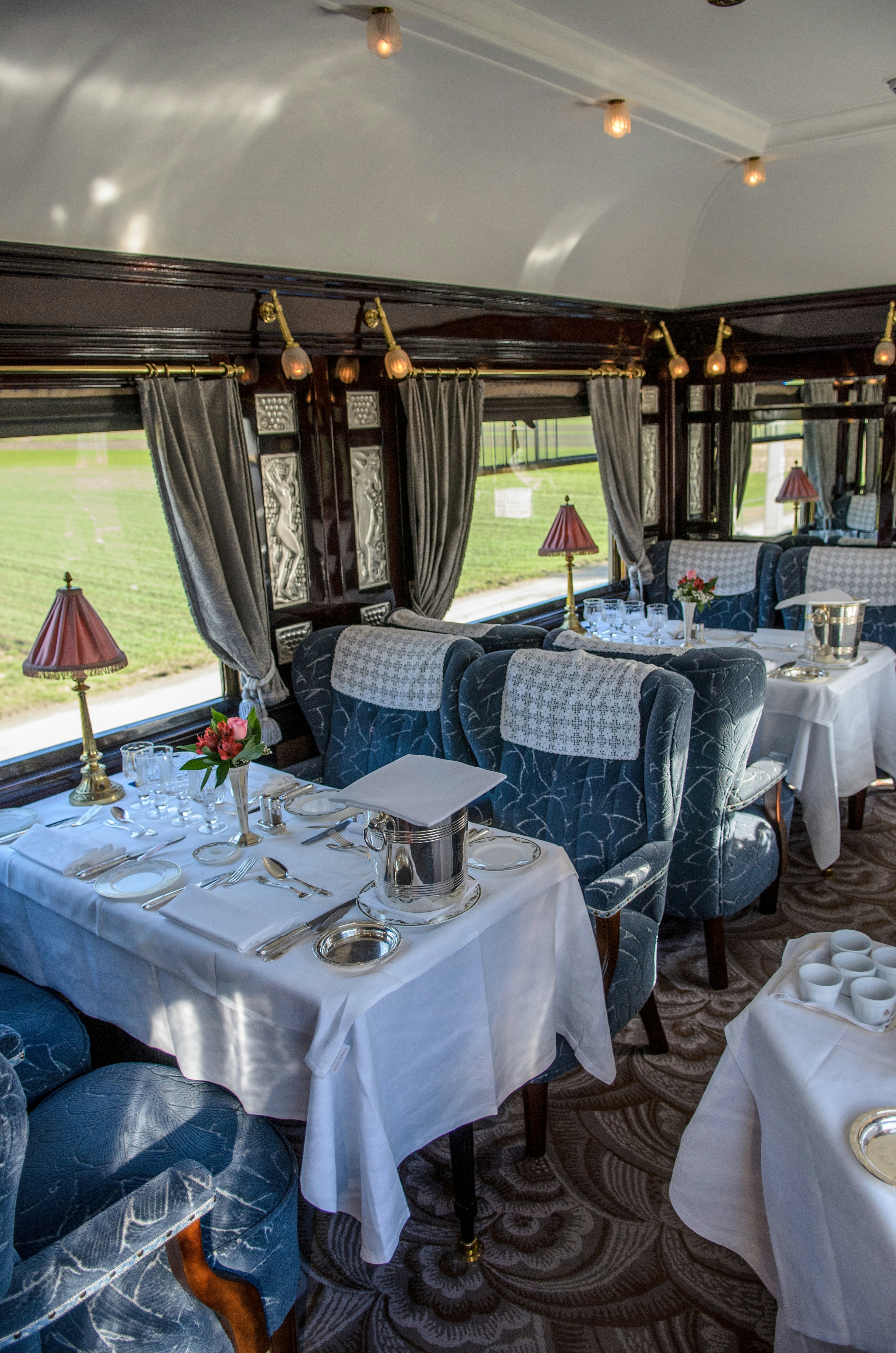 Atmospheric dining car interior with white linen tables, crystal glassware, brass wall sconces, and panoramic windows showing twilight landscape
