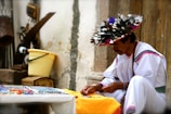 A person wearing traditional attire is seated outdoors, focusing intently on a craft activity. The outfit is predominantly white with colorful accents and a headdress adorned with feathers. Various craft items are placed on a table nearby, and there are rustic tools and a yellow bucket against a weathered wall.