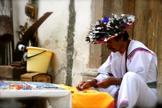 A person wearing traditional attire is seated outdoors, focusing intently on a craft activity. The outfit is predominantly white with colorful accents and a headdress adorned with feathers. Various craft items are placed on a table nearby, and there are rustic tools and a yellow bucket against a weathered wall.