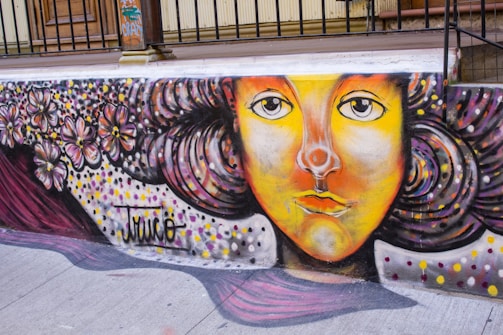 A vibrant street mural features a stylized face with expressive eyes and a mix of orange, yellow, and red hues on the skin. The hair is depicted with swirling patterns in dark colors and is adorned with pink and purple flowers. The mural includes multicolored dots, adding to its whimsical appearance, and the name 'Truco' is signed on the artwork.