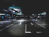 A dimly lit parking garage with a concrete floor, illuminated by ceiling lights. White arrows and parking lines are painted on the ground, directing traffic flow. The environment appears empty and vast, with support beams visible.