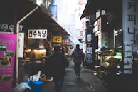 A bustling alleyway in a Korean market filled with people walking among various food stalls and shops. Brightly colored signs in Korean hang above the stalls, and there is a mix of smoke or steam in the air, suggesting that food is being cooked. The environment seems lively and busy, with people engaged in different activities, from shopping to preparing food.