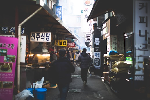 A bustling alleyway in a Korean market filled with people walking among various food stalls and shops. Brightly colored signs in Korean hang above the stalls, and there is a mix of smoke or steam in the air, suggesting that food is being cooked. The environment seems lively and busy, with people engaged in different activities, from shopping to preparing food.