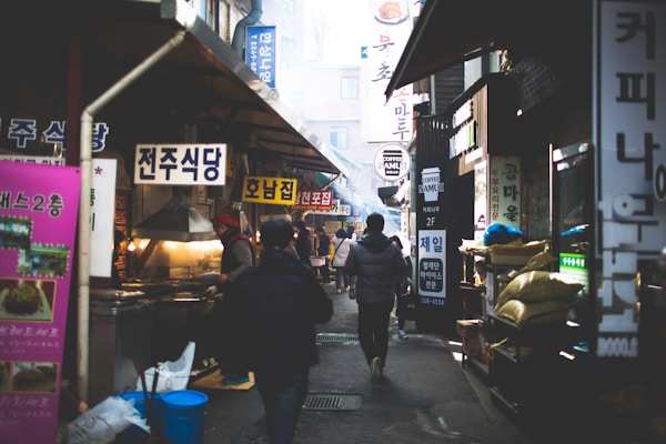 A bustling alleyway in a Korean market filled with people walking among various food stalls and shops. Brightly colored signs in Korean hang above the stalls, and there is a mix of smoke or steam in the air, suggesting that food is being cooked. The environment seems lively and busy, with people engaged in different activities, from shopping to preparing food.
