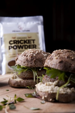 A close-up view of two burgers made with dark buns and topped with fresh greens. The background shows a bag labeled 'Cricket Powder,' indicating the unique ingredient used in the preparation of the burgers. The buns are sprinkled with sesame seeds.