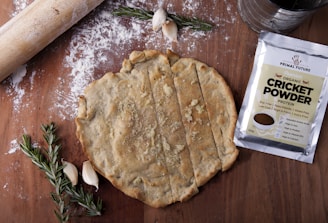 A wooden surface with a rolled-out dough, partially sliced into rectangles. Next to the dough, a packet labeled 'Primal Future Organic Cricket Powder' is visible. A rolling pin, scattered flour, garlic cloves, and sprigs of rosemary are also present.