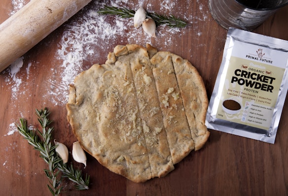 A wooden surface with a rolled-out dough, partially sliced into rectangles. Next to the dough, a packet labeled 'Primal Future Organic Cricket Powder' is visible. A rolling pin, scattered flour, garlic cloves, and sprigs of rosemary are also present.
