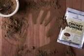 A wooden surface is covered with a light brown powder, with a handprint visible in the scattered substance. A small white bowl filled with the same powder is accompanied by a metal spoon. Nearby, a packet labeled 'Organic Cricket Powder' displays various nutrition labels and claims about the product, such as being soy-free and protein-rich.